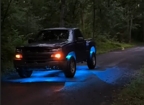 Lifted Chevy Silverado at dusk on a wooded trail with bright blue underglow rock lights illuminating the ground